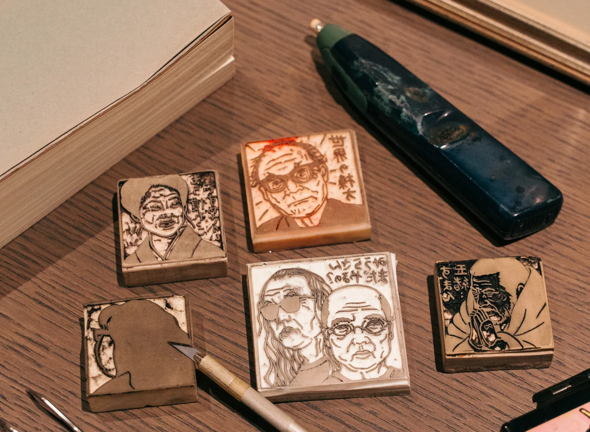 浅虫温泉 椿館の「ナンシー関 展示」。木目調の机に並ぶ、石原慎太郎やいとうせいこうを描いた消しゴム版画の原版。制作道具のカッターと共に、手仕事の凄みと創作の息吹を間近に感じさせる展示カット