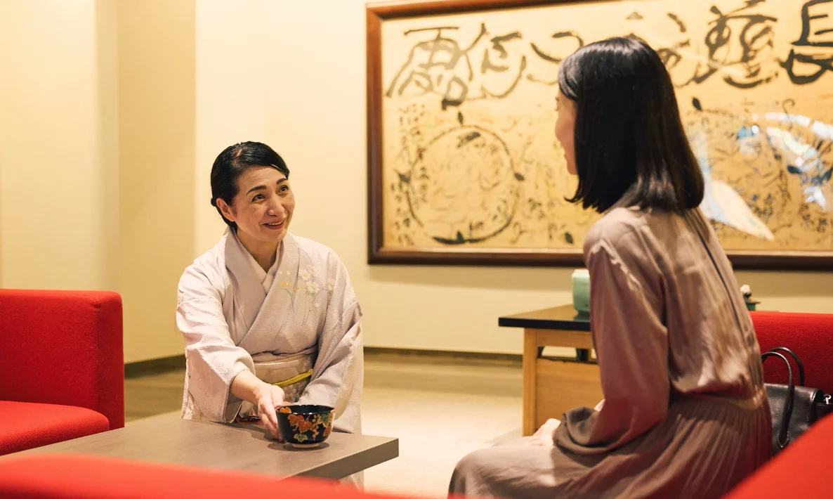 浅虫温泉 椿館の「棟方志功ギャラリー」で、巨匠の力強い作品を背景に、女将の温かなおもてなしを受けるひととき。アートに囲まれたモダンな赤いソファで楽しむお茶の時間が、心豊かな滞在を演出する。歴史ある芸術作品と、今に息づく真心が交差する椿館ならではの贅沢な風景